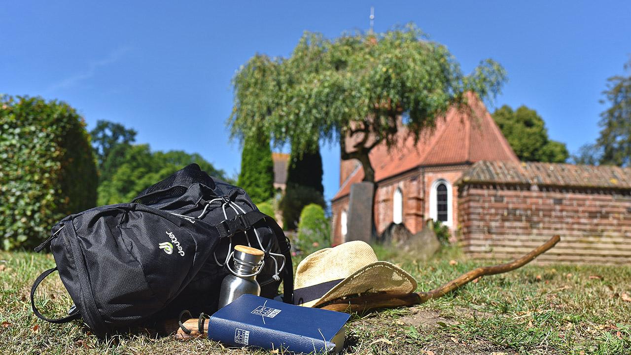 Ein Rucksack, ein Hut und eine Trinkflasche liegen auf dem Gras vor einer kleinen Kirche