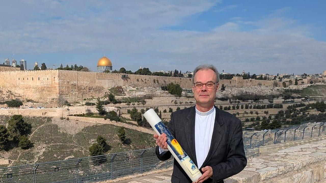 Ein Mann im dunklen Anzug trägt eine große Kerze vor der Stadtansicht Jerusalems mit Felsendom