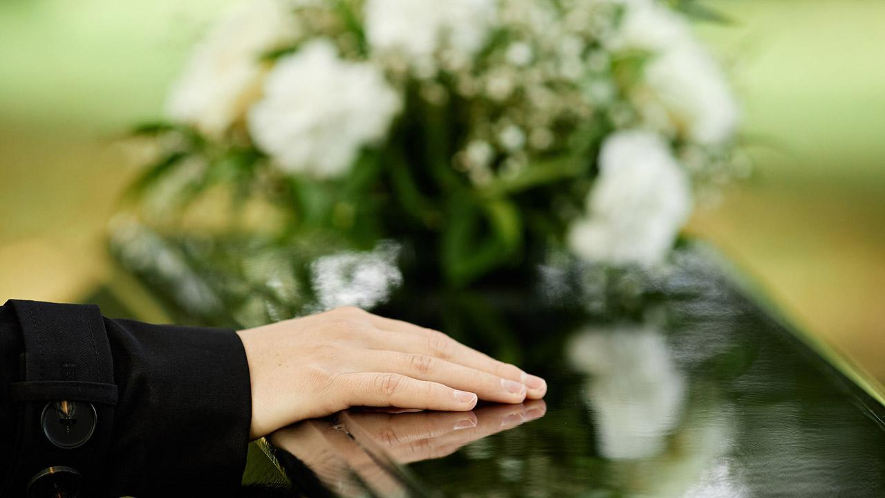 Eine Hand und ein weißes Blumengesteck auf einem schwarzen Sarg