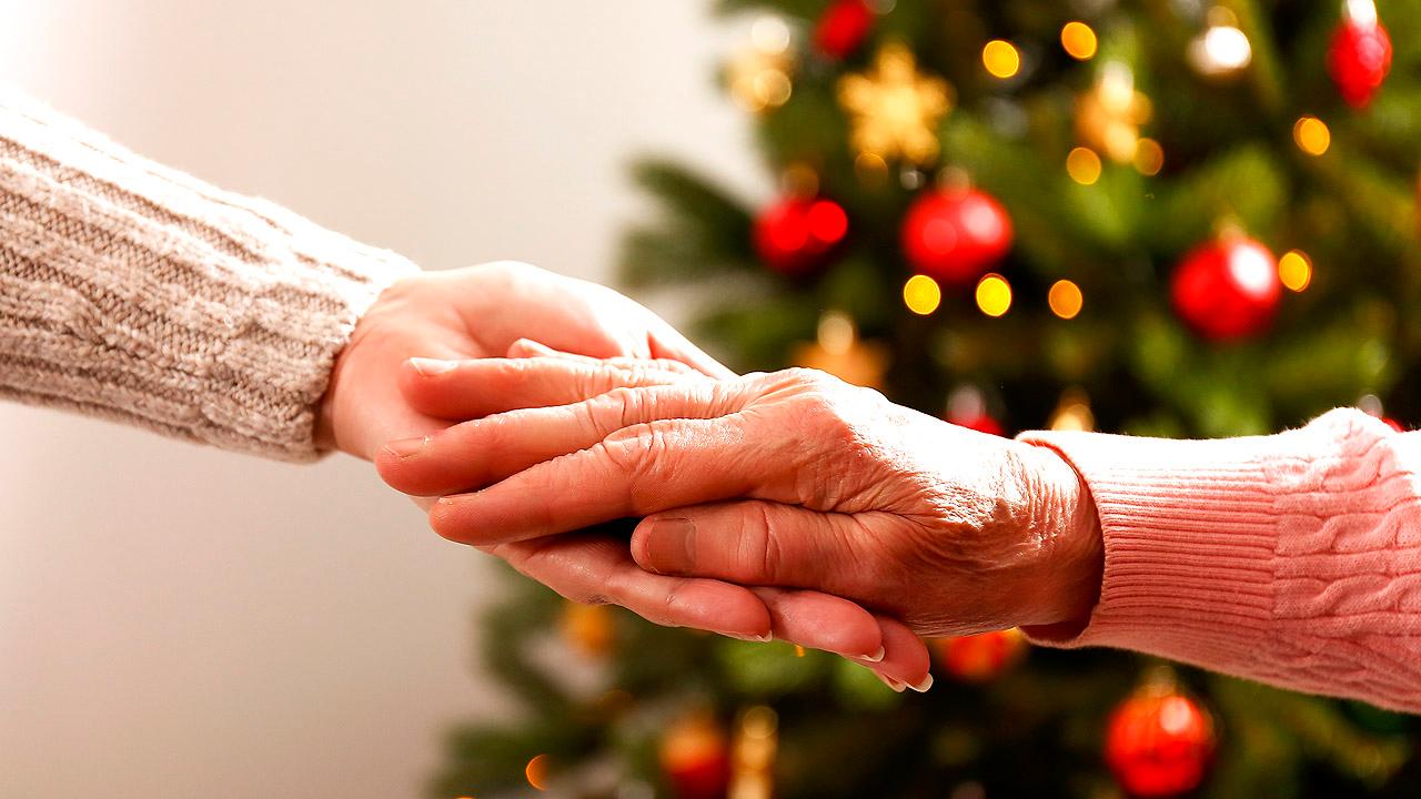 Eine Hand hält die Hand einer älteren Dame, dahinter ein geschmückter Weihnachtsbaum