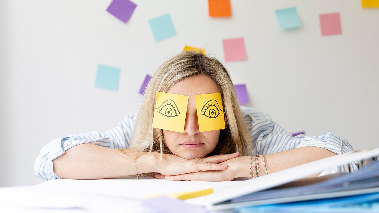 Eine Frau stützt ihren Kopf im Büro auf ihre Hände ab, auf ihren geschlossenen Augen kleben Post-It-Zettel mit aufgemalten wachen, offenen Augen