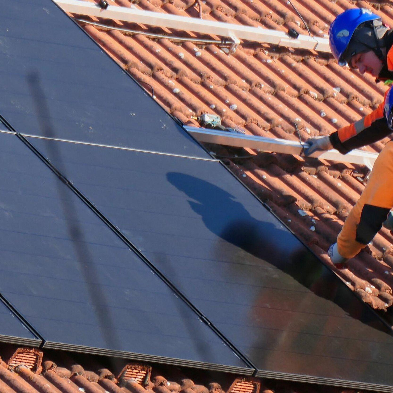 Zwei Handwerker installieren eine Solaranlage auf einem Hausdach. Die Handwerker tragen Bereufskleidung und Helme zur Sicherheit.