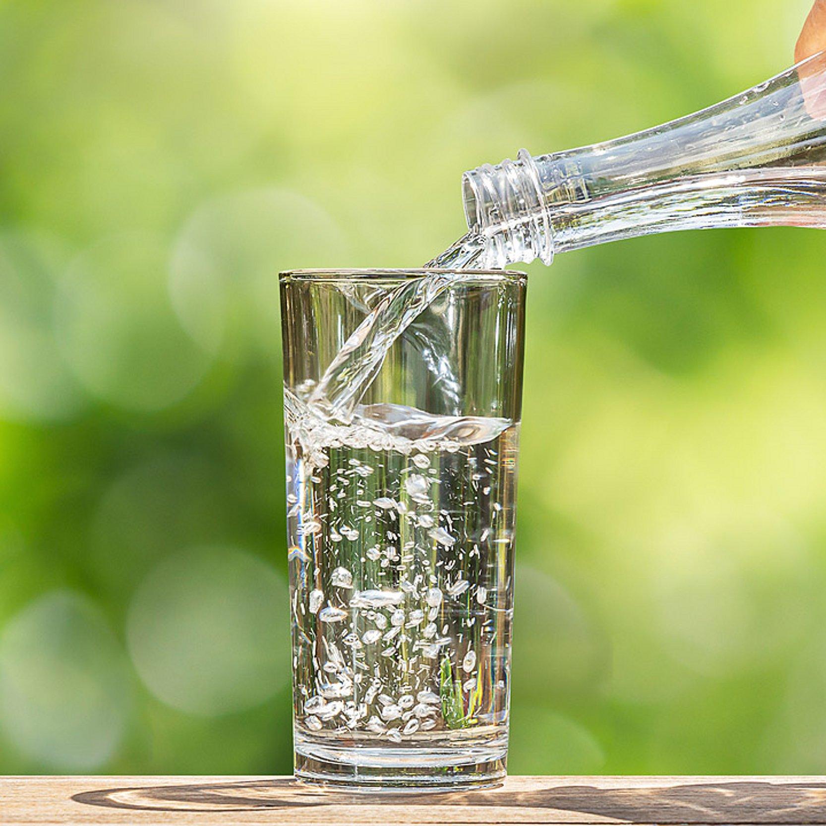 Eine Hand hält eine Flasche Wasser, aus der Wasser in ein Glas fließt. Der Hintergrund ist grün.