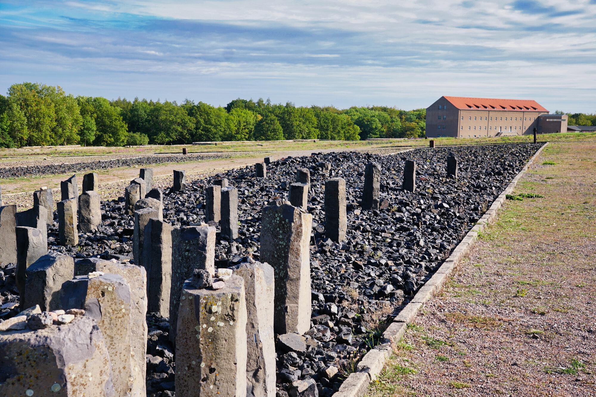 Gedenken an die Sinti und Roma;  KZ Gedenkstätte Buchenwald. Gedenksteine im Bildvordergrund. Im Hintergrund ein Gebäude, am rechten Bildrand Bäume und oben blauer Himmel mit Wolken.