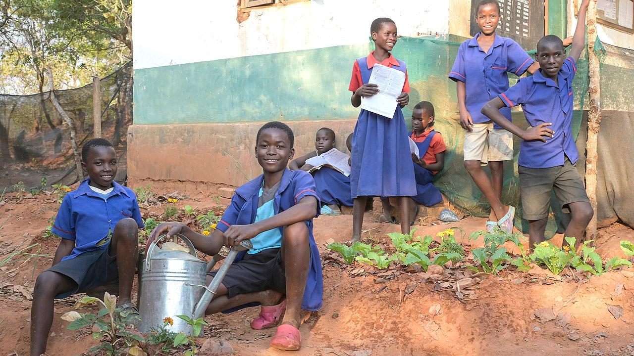 Afrikanische Schüler sind mit einer Gießkanne im Schulgarten, ein Mädchen hat ein Heft in der Hand