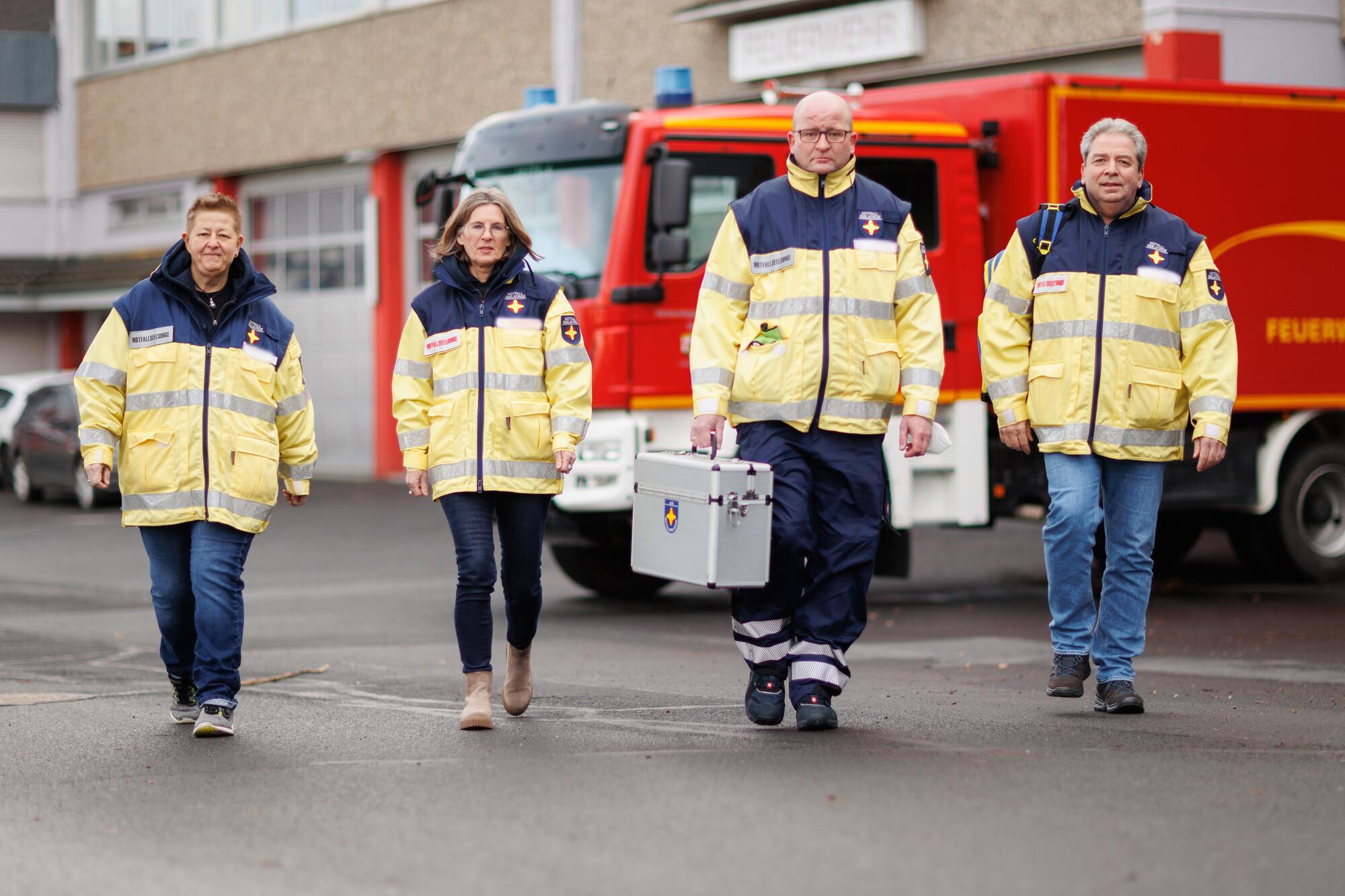 Vier Personen laufen auf den Fotografen zu. Sie tragen Uniformen der Notfallseelsorge. Im Hintergrund steht ein Feuerwehrfahrzeug vor der Brandwache.