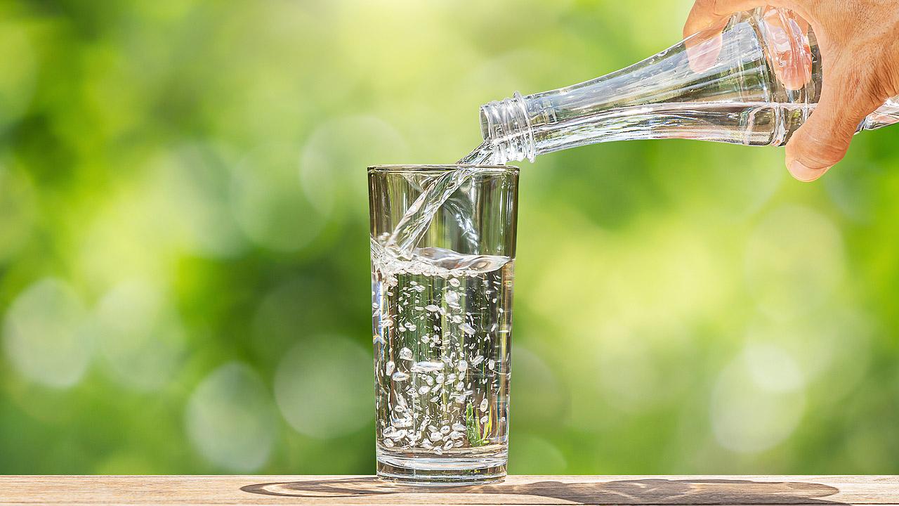 Eine Hand hält eine Flasche Wasser, aus der Wasser in ein Glas fließt. Der Hintergrund ist grün.