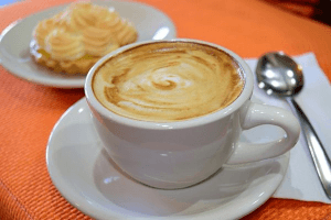 Weiße Porzellantasse mit Kaffee und Milchschaum, dahinter ein kleiner Teller mit Gebäck