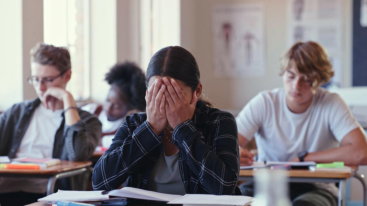 Prüfungssituation in der Schule, eine Person im Vordergrund hält sich verzweifelt die Hände vor das Gesicht