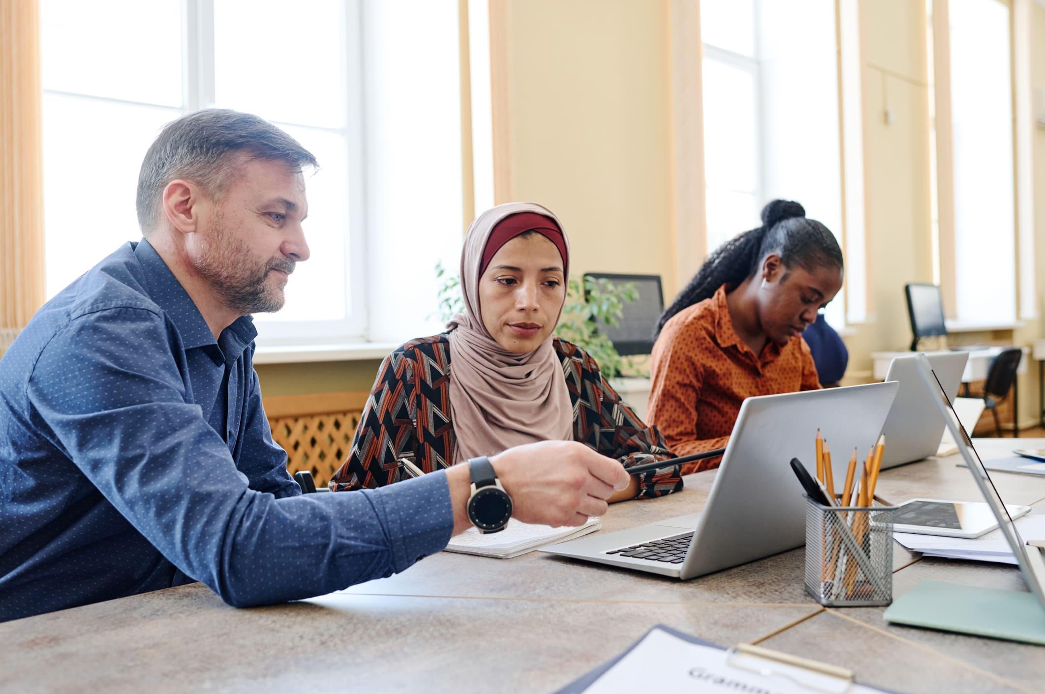 Ein Mann erklärt einer Frau mit Migrationshintergrund eine Aufgabe an einem Computer