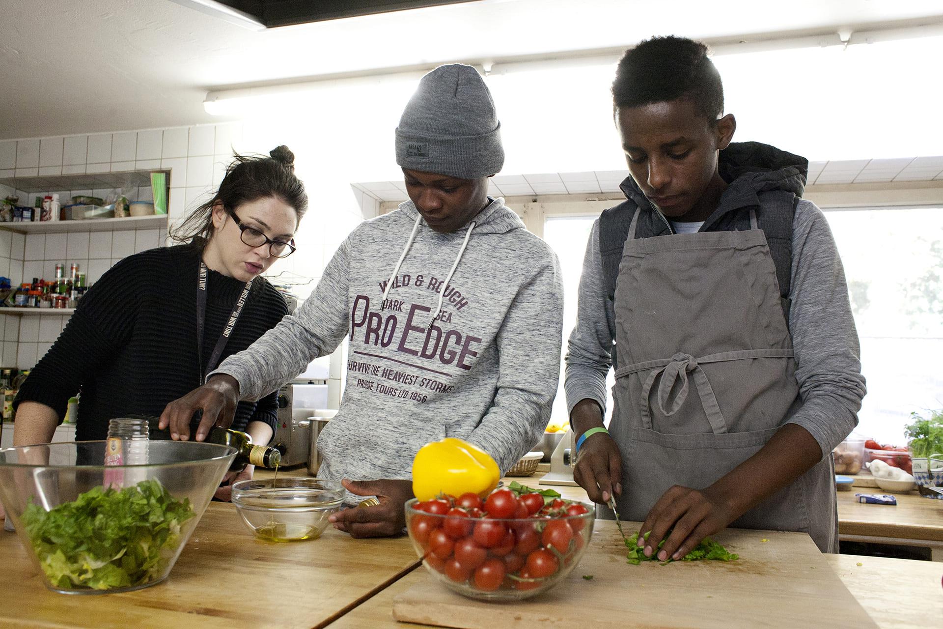 Flüchtlinge kochen mit junger Frau Zwei junge Geflüchtete kochen gemeinsam mit einer jungen Frau im Vordergrund steht eine Schüssel mit Tomaten.
