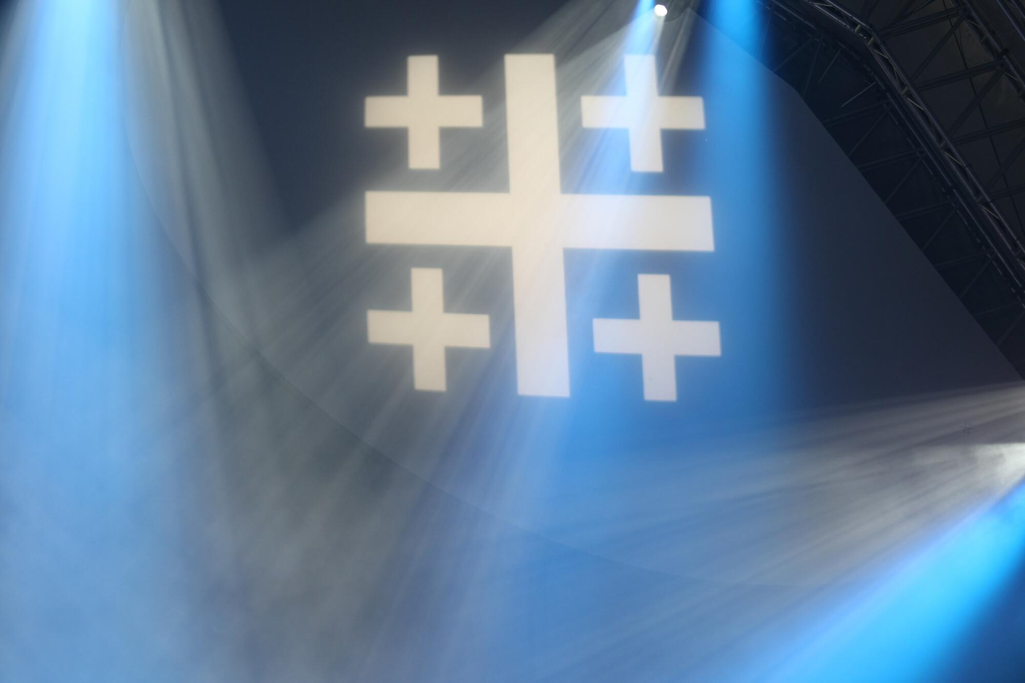 Ein Jerusalemkreuz wird in einer blauen Halle projiziert und Scheinwerfer bestrahlen es