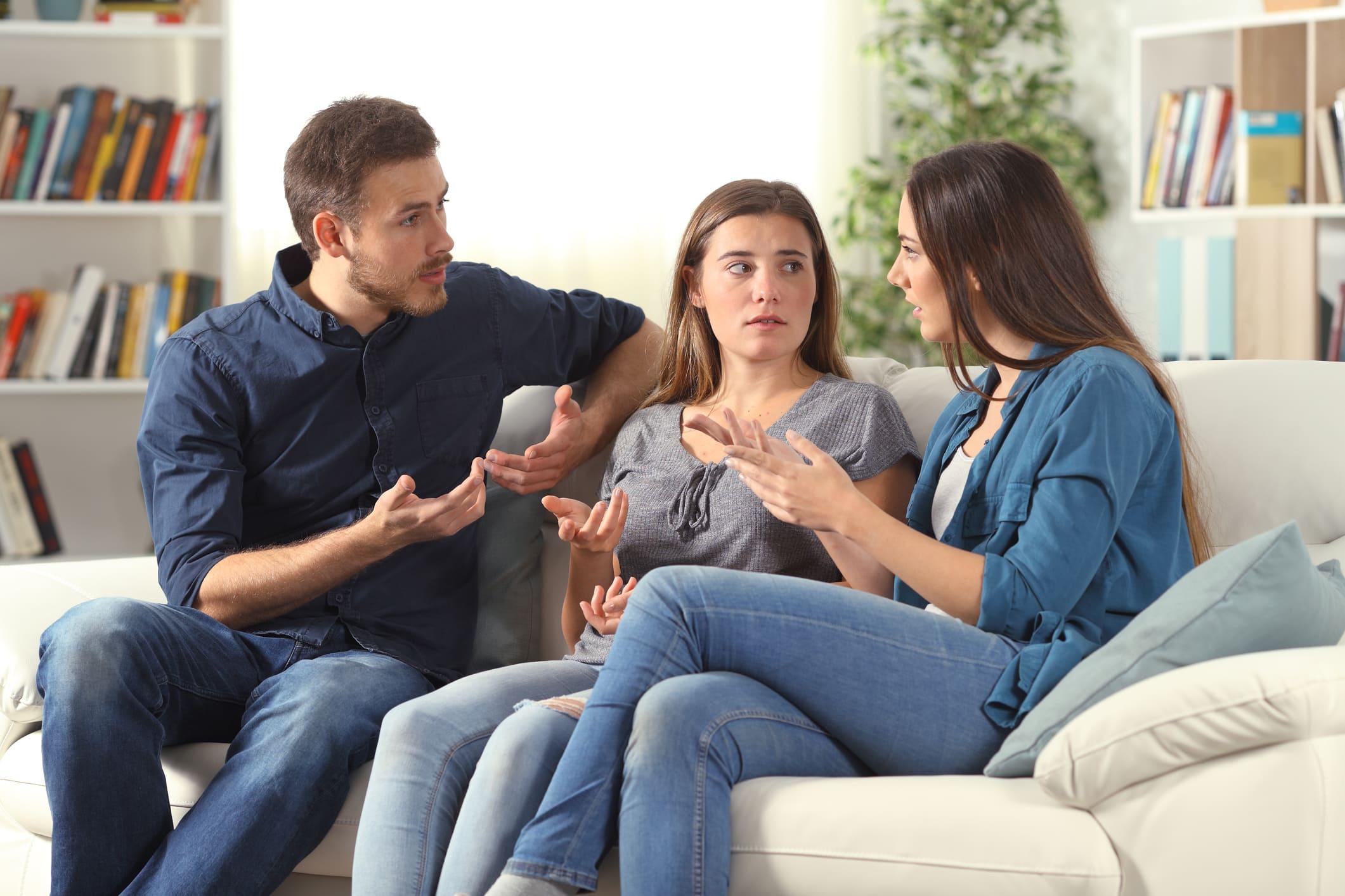 Ein junger Mann und zwei junge Frauen sitzen auf einer weißen Couch und diskutieren lebhaft miteinander, sie gestikulieren dabei.