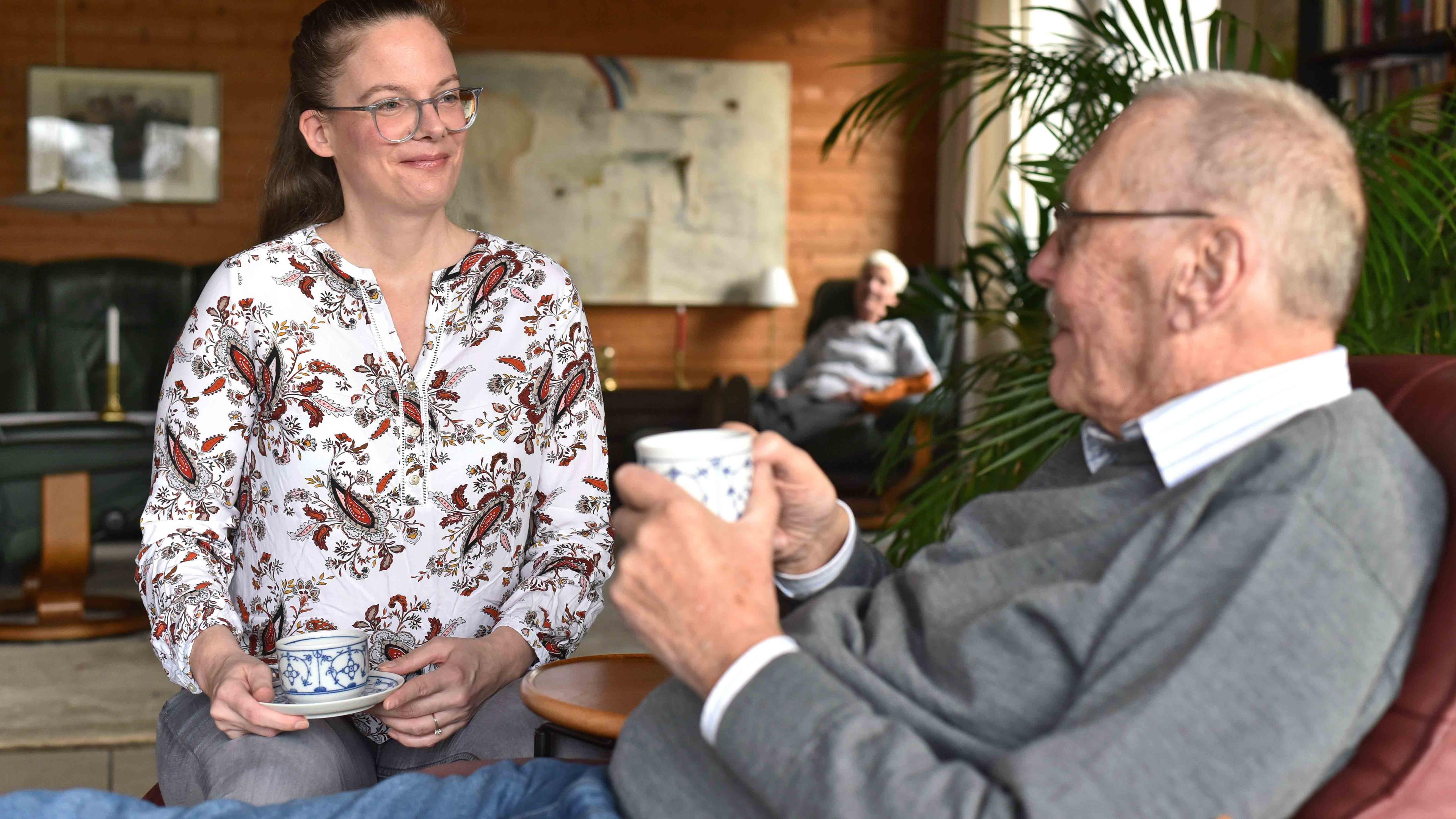 Eine Frau und ein älterer Mann sitzen sich bei einer Tasse Kaffee gegenüber und unterhalten sich in einem gemütlichen Wohnzimmer, im Hintergrund sitzt eine Frau.