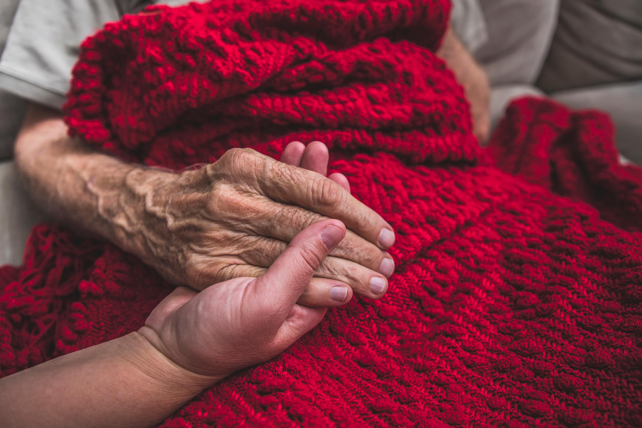 Eine junge Hand hält die alte Hand einer Sterbenden. Diese ist mit einer schönen, roten Decke zugedeckt.