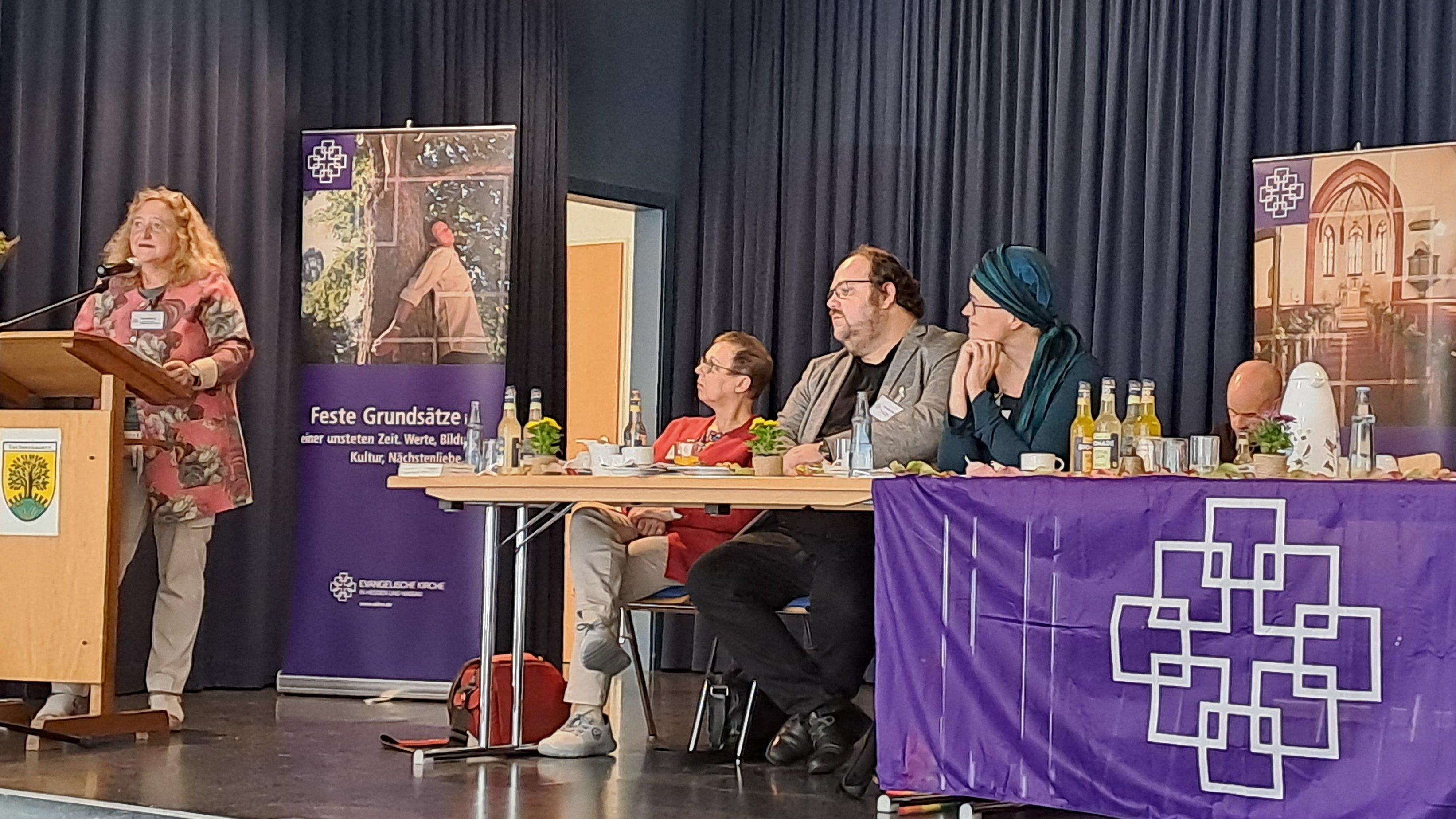 Auf einem Podium steht links eine Frau am Rednerpult. Rechts sitzen mehrere Personen hinter einem Tisch Das Podium ist geschmückt mit lila Banner der Evangelischen Kirche in Hessen und Nassau.