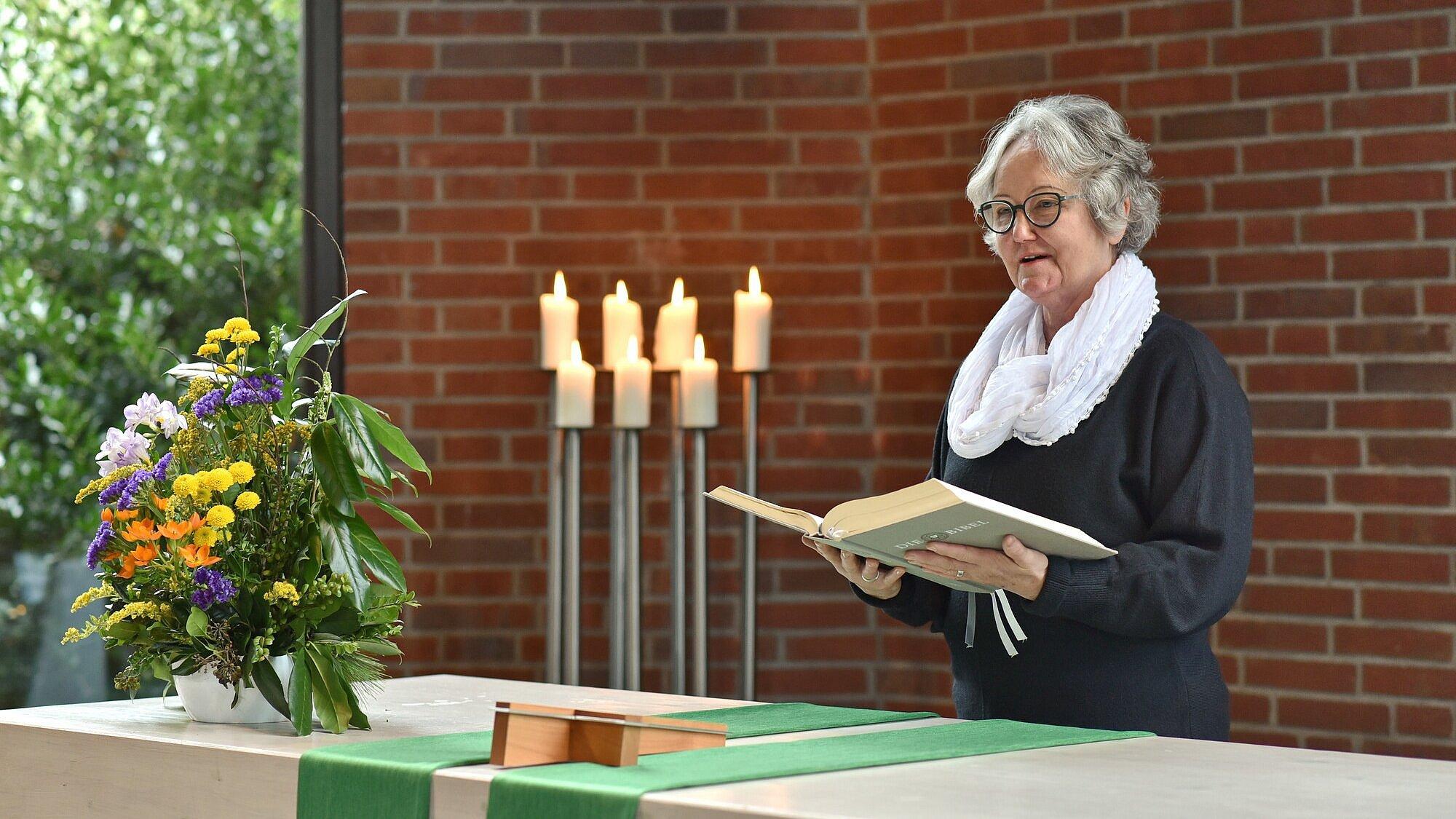 Ein predigende Frau steht hinter einem Altar mit Blumen und liest aus einer großen Bibel, die sie in Händen hält. Im Hintergrund brennen sieben Kerzen auf hohen silbernen Lauchtern.