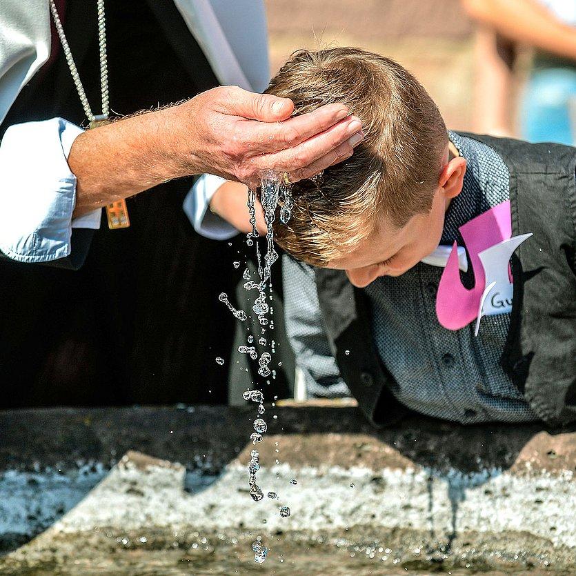 Ein Junge im Anzug beugt sich vor und wird getauft, durch die Hand eines Pfarrers fließt Wasser über seine Stirn