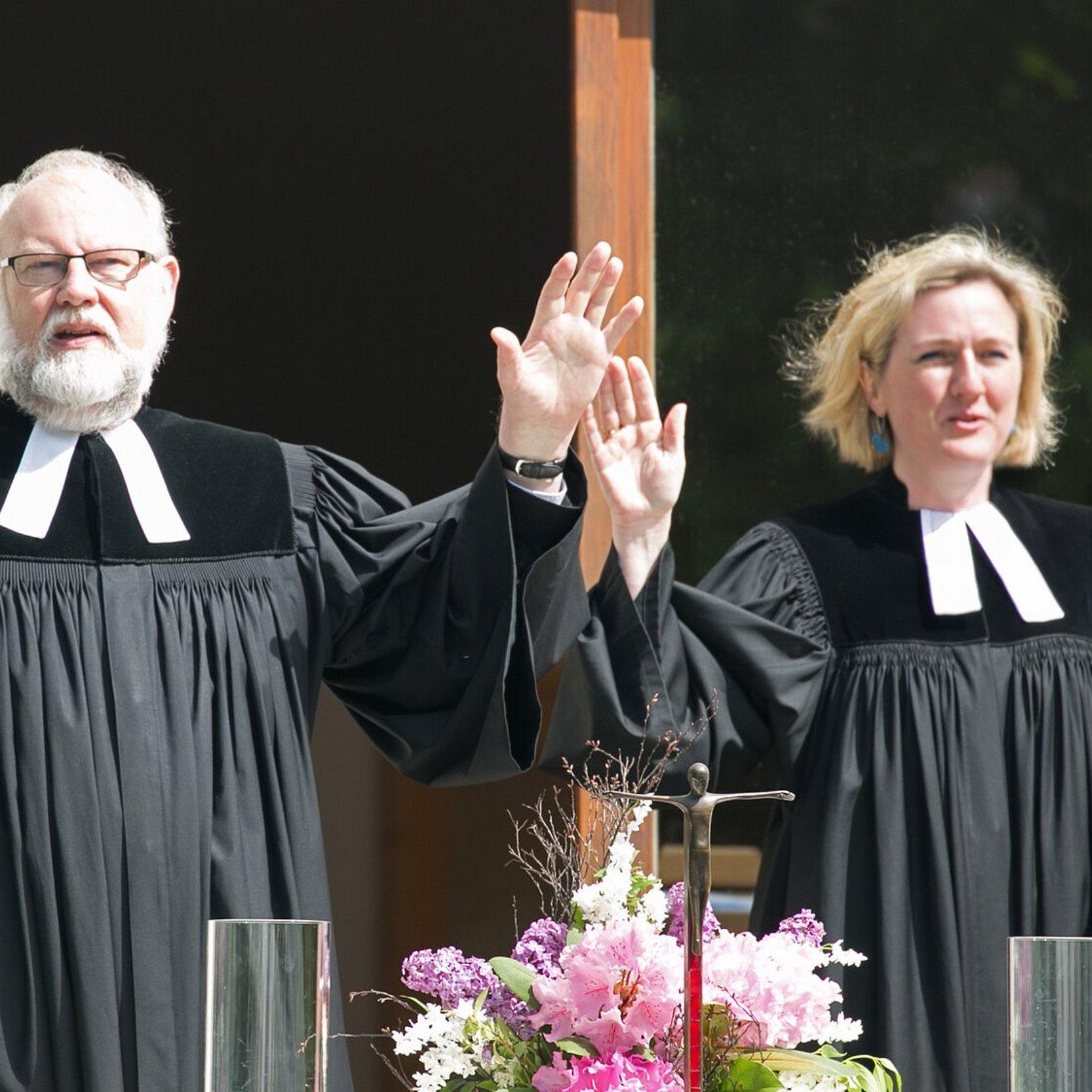 Ein Pfarrer und eine Pfarrerin erheben die Hände und segnen. Das Foto zeigt grüne Umgebung, Garten, Altar mit Kerzen, Kreuz und Blumen