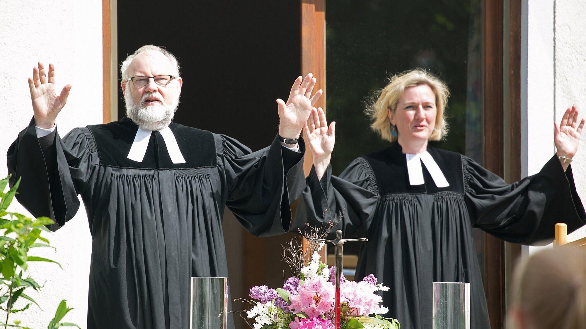 Ein Pfarrer und eine Pfarrerin erheben die Hände und segnen. Das Foto zeigt grüne Umgebung, Garten, Altar mit Kerzen, Kreuz und Blumen