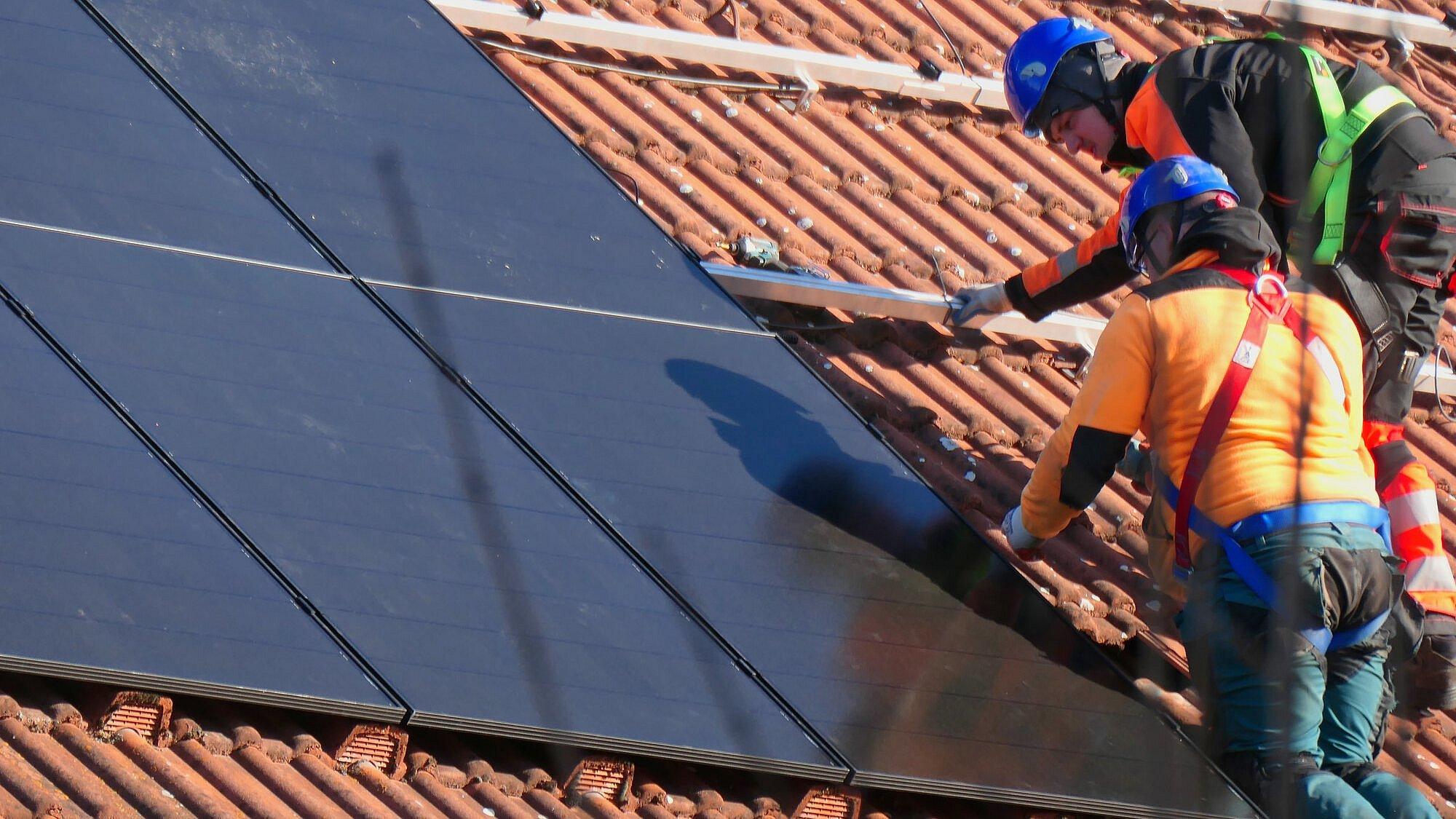 Zwei Handwerker installieren eine Solaranlage auf einem Hausdach. Die Handwerker tragen Bereufskleidung und Helme zur Sicherheit.