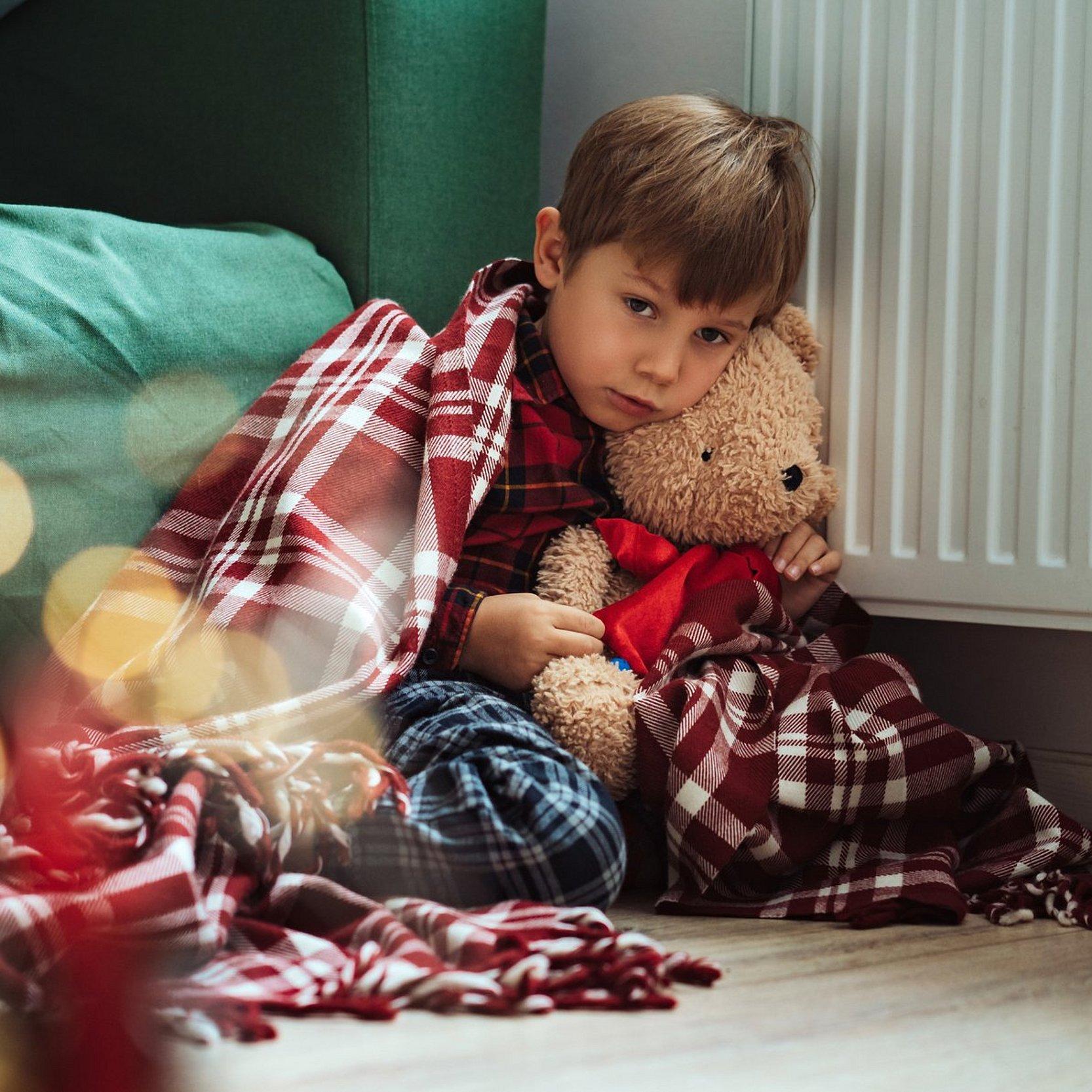 Wärmewinter Frierendes Kind sitzt mit Teddy auf dem Boden an der Heizung.