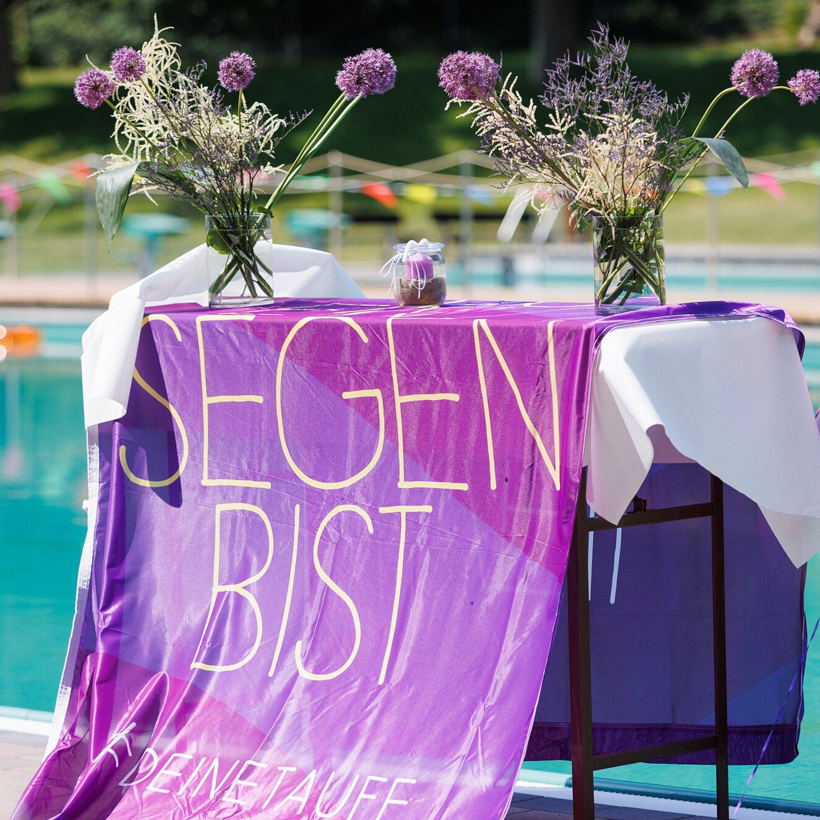 Tauffest im Naturschwimmbad Höhr-Grenzhausen_004 Tauffest im Naturschwimmbad Höhr-Grenzhausen: Eine Fahne der Taufinitiative der EKD liegt auf einem Tisch und hängt bis zum Boden. Der Tisch steht vor einem Schwimmbecken und ist mit Blumen und Taufkerzen dekoriert. Aufnahme seitlich von rechts.