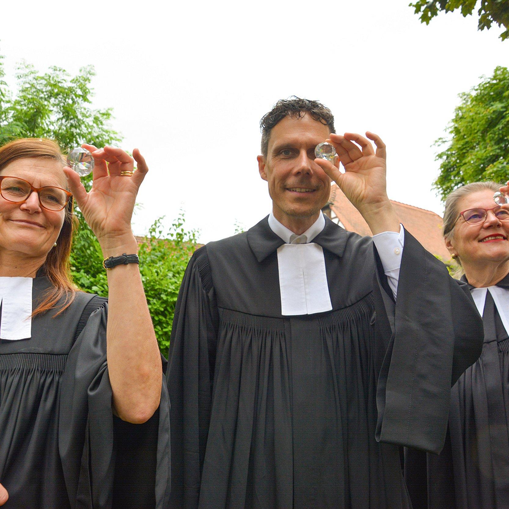 Pfarrteam Ein Pfarrer und zwei Pfarrerinnen halten sich einen Kristall im Facettschliff vor Augen