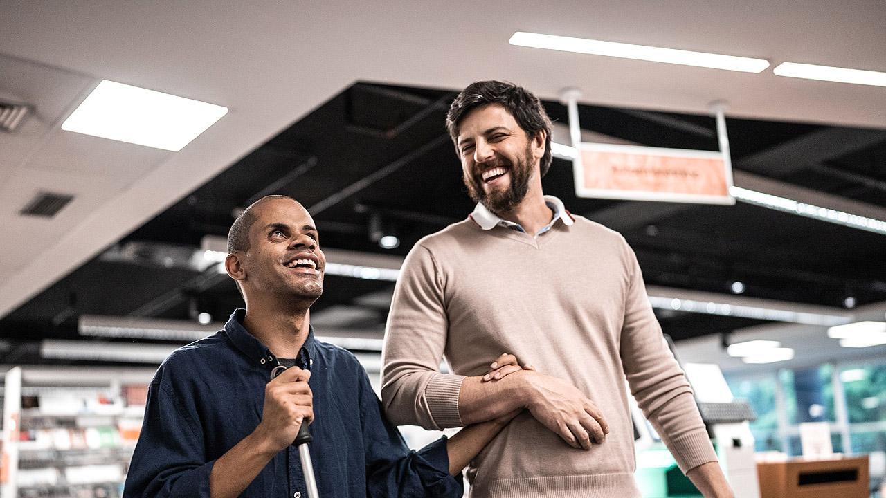 Inklusion Ein Mann führt einen blinden Mann durch eine Bibliothek