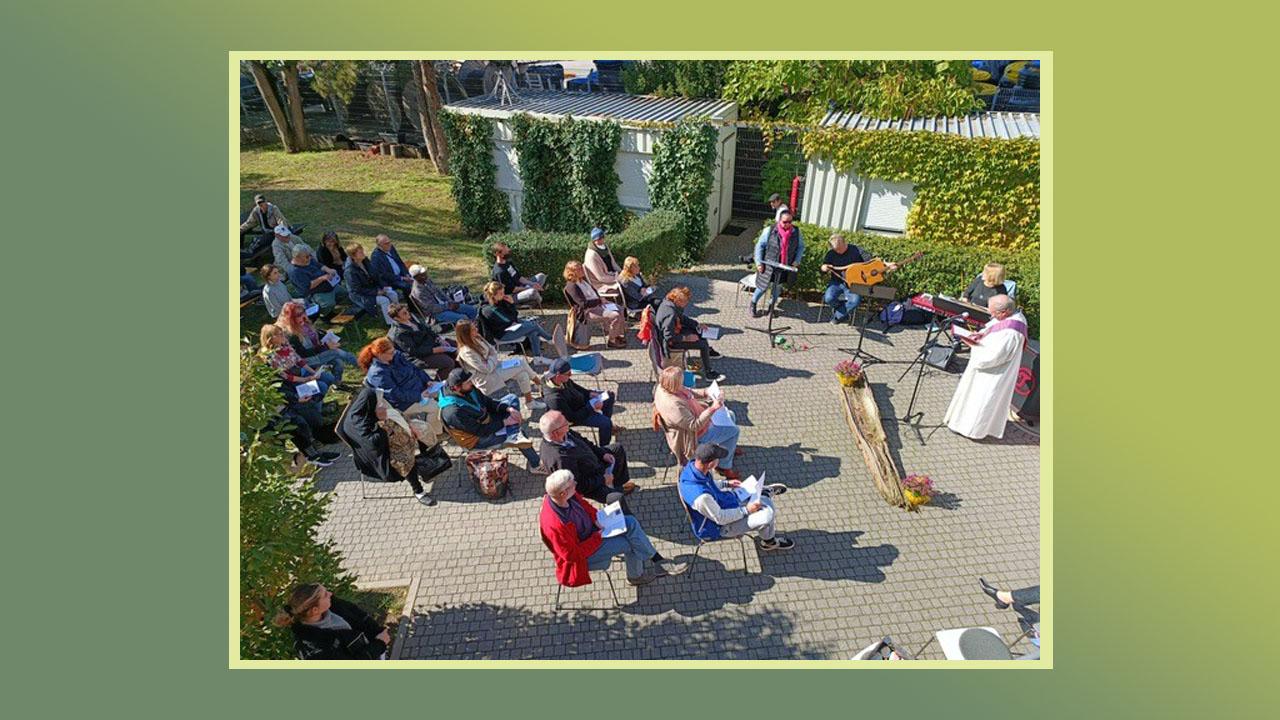 Gedenkgottesdienst Menschen sitzen draußen im Grünen, vorne stehen ein Pfarrer im schwarzen und einer im weißen Talar