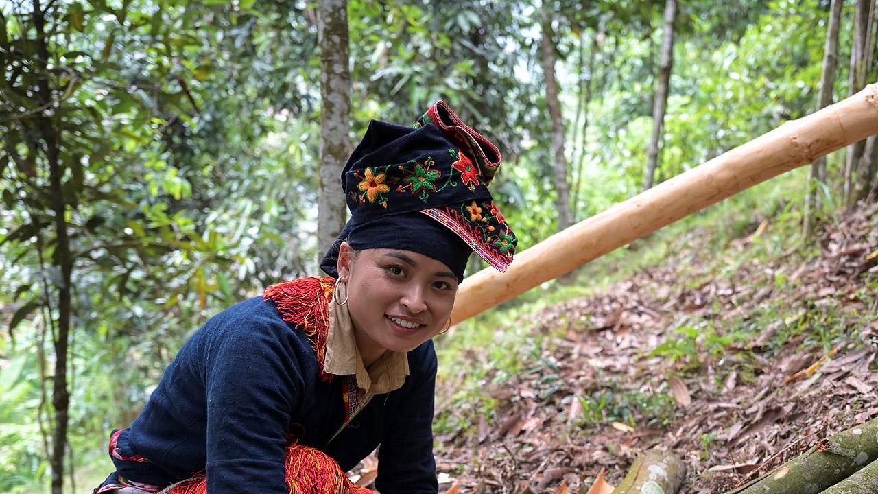 Frau mit wertvoller Tracht kniet neben Baumrindenstücken in einem Wald