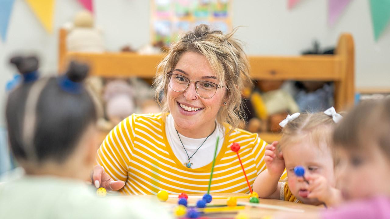 Eine Erzieherin mit gelbem Pullover in einer Kita, vor ihr drei Kinder