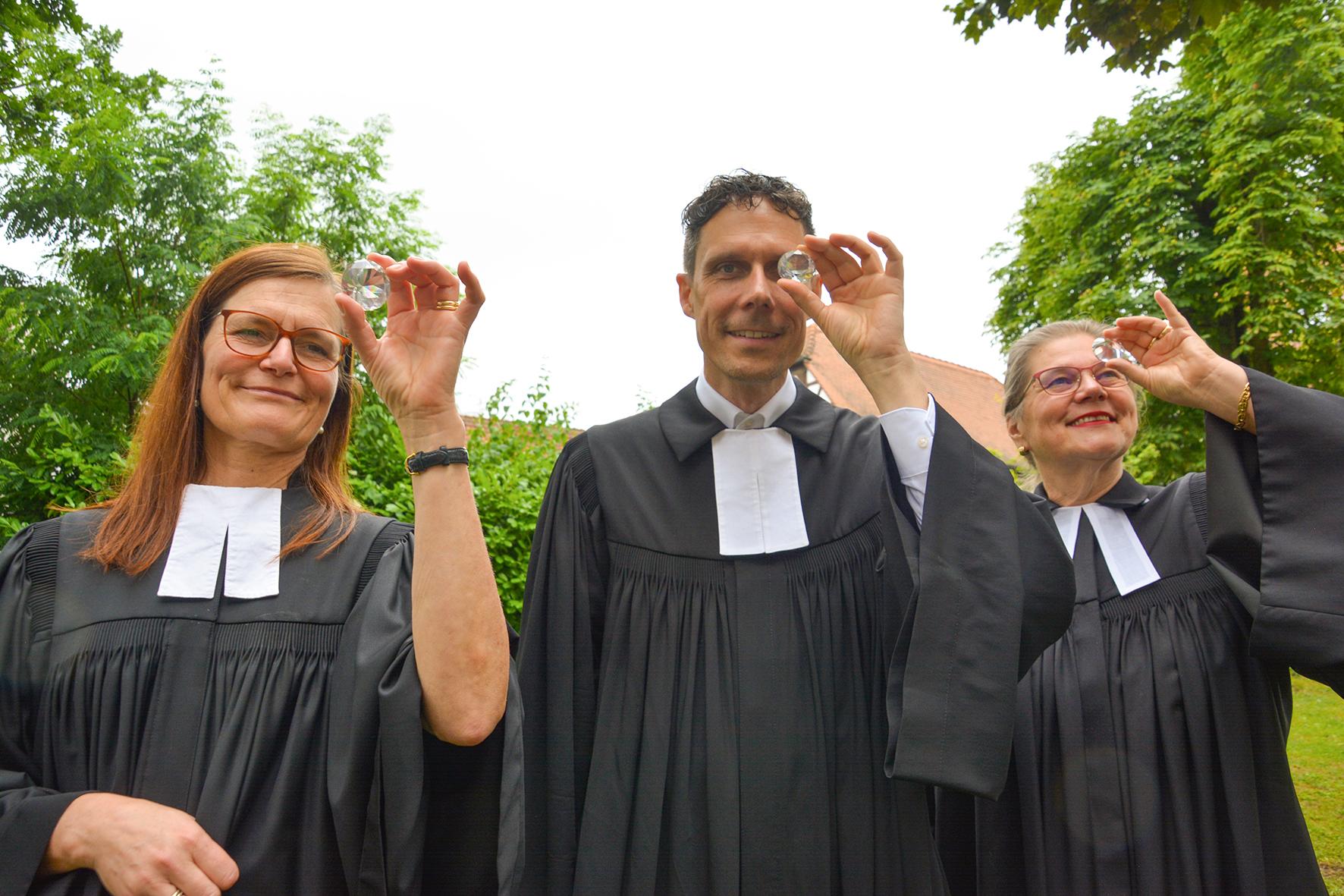 Ein Pfarrer und zwei Pfarrerinnen halten sich einen Kristall im Facettschliff vor Augen