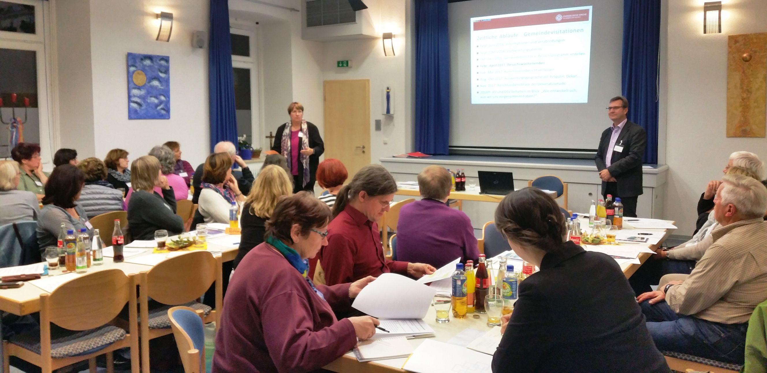 Menschen an Konferenztischen, im HIntergrund Präsentation auf Leinwand
