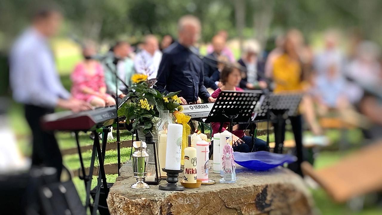 Ein Tauffest findet draußen statt. Auf einem Fels stehen mehrere Taufkerzen im Hintergrund spielt eine Band Musik.