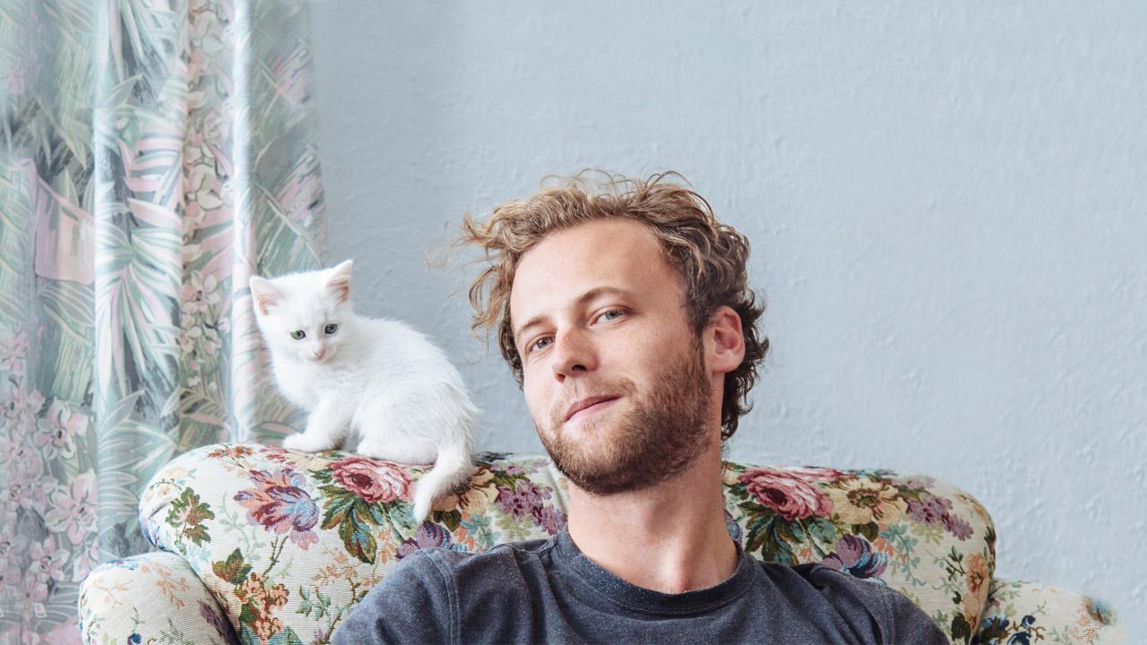 Junger Mann sitzt auf einem Sessel mit Blumenmuster, auf der Lehne sitzt eine junge, weiße Katze