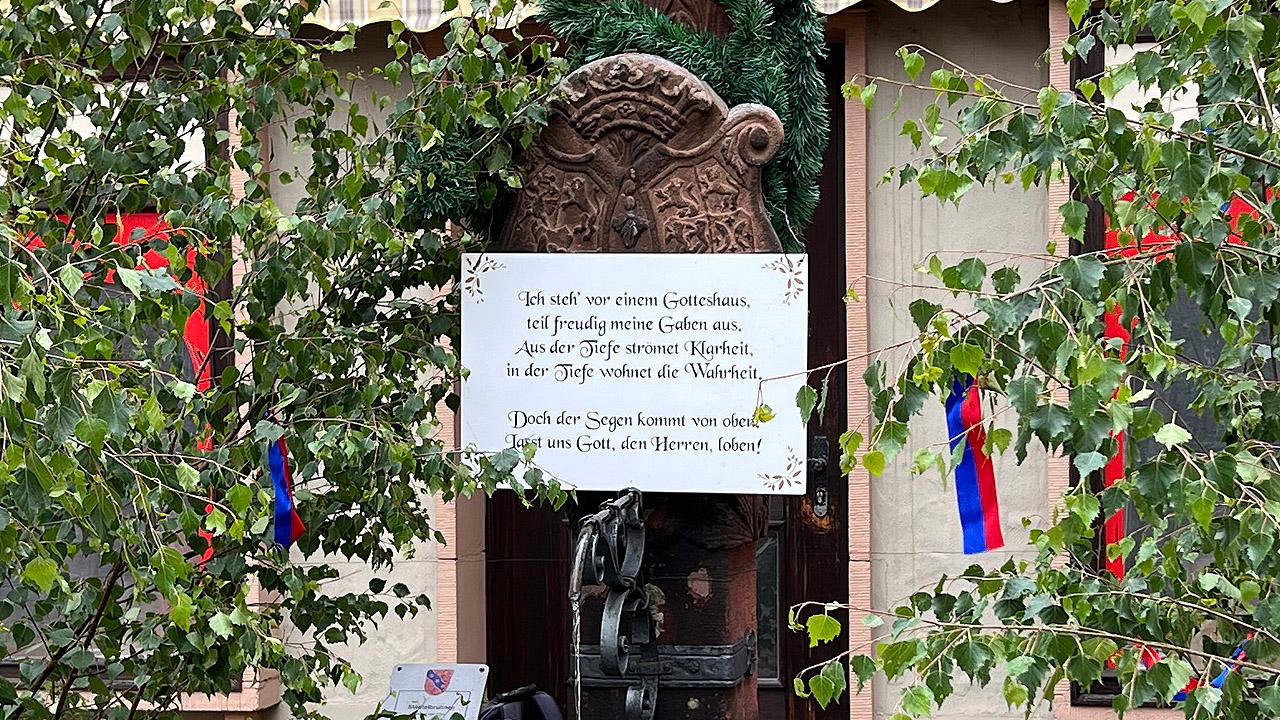 Birkengrün mit Bändern um einen Brunnen mit dem Schild: Ich steh vor einem Gotteshaus, teil freudig meine Gaben aus. Aus der Tiefe strömet Klarheit, in der Tiefe wohnet die Wahrheit. Doch der Segen kommt von oben. Lasst uns Gott, den Herrn loben!