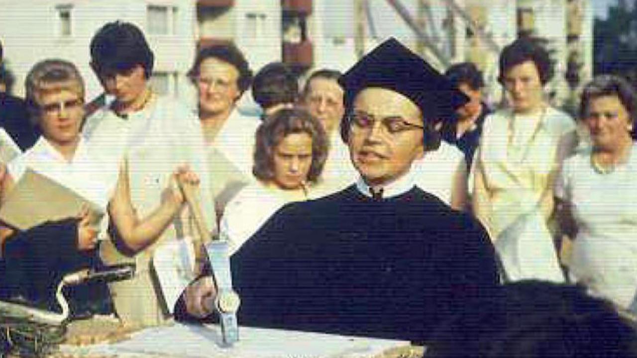 Pfarrerin im alten schwarzen Talar mit schwarzer Haube haut mit einem Hammer auf einen Stein im Hintergrund stehen Menschen im Halbkreis und schauen ihr zu.