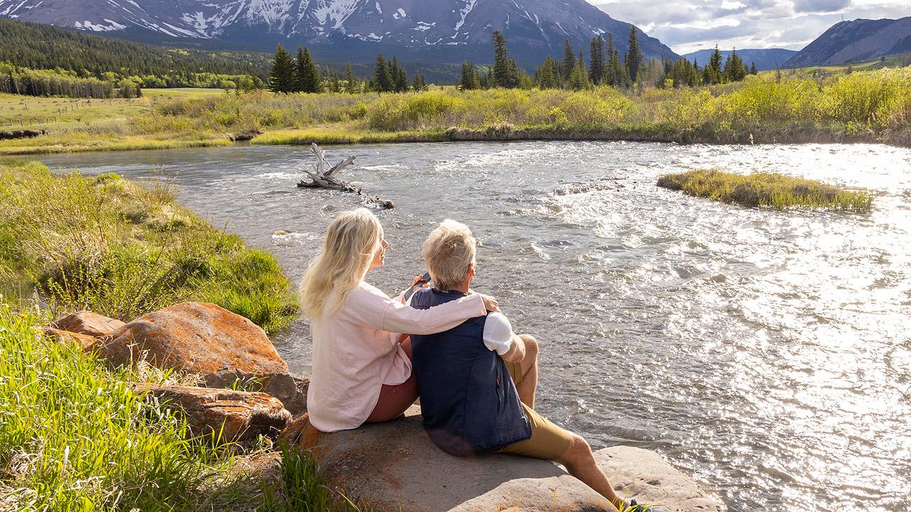 Ein älteres Paar sitzt an einem Fluss in einer alpinen Landschaft, die Frau hat die Hand auf die Schulter des Mannes gelegt