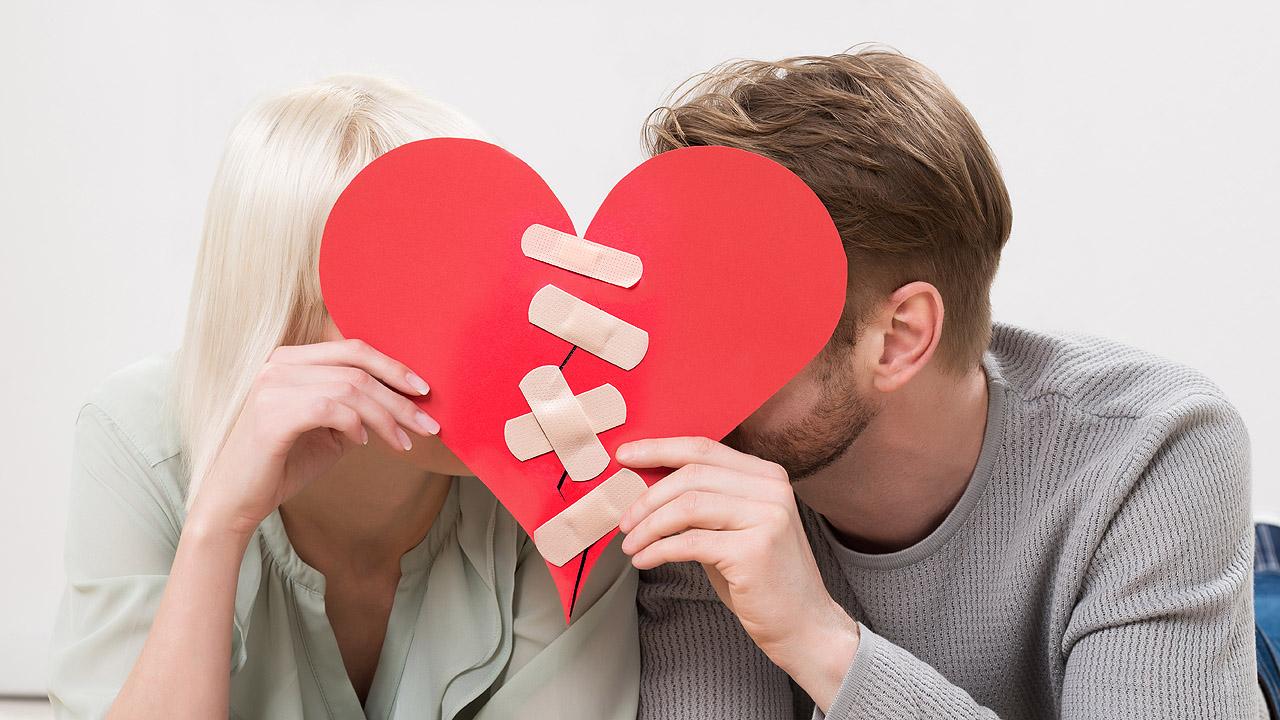 Paar hinter einem roten Pappherz, durch das ein Riss geht, der mit Wundpflastern zugeklebt wurde