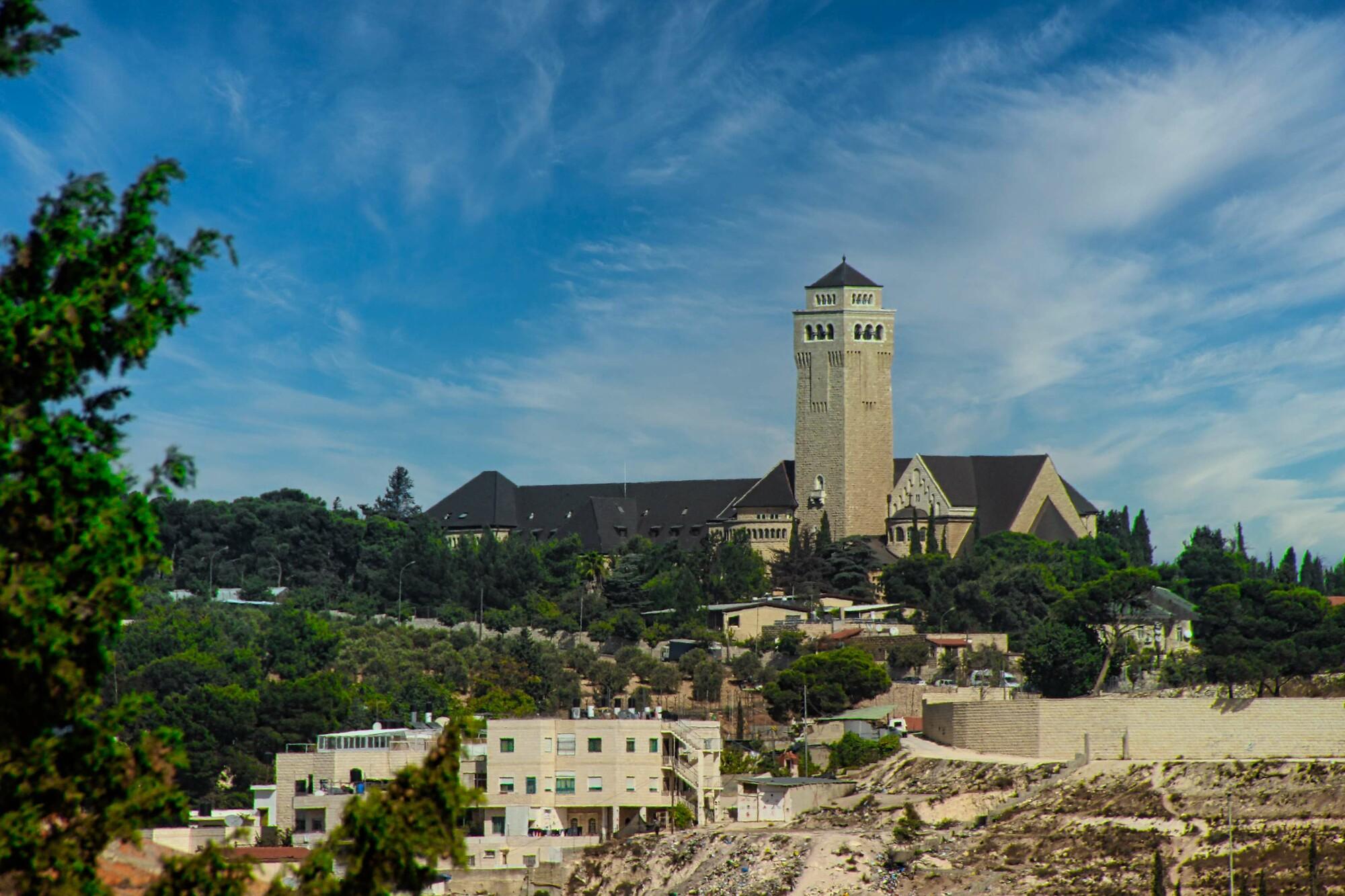 Auguste Victoria Kirche auf dem Ölberg in Jerusalem Große Kirche mit hohem Turm auf einem Hügel, umgeben von Bäumen und Wohnhäusern, unter einem blauen Himmel mit feinen Wolken.