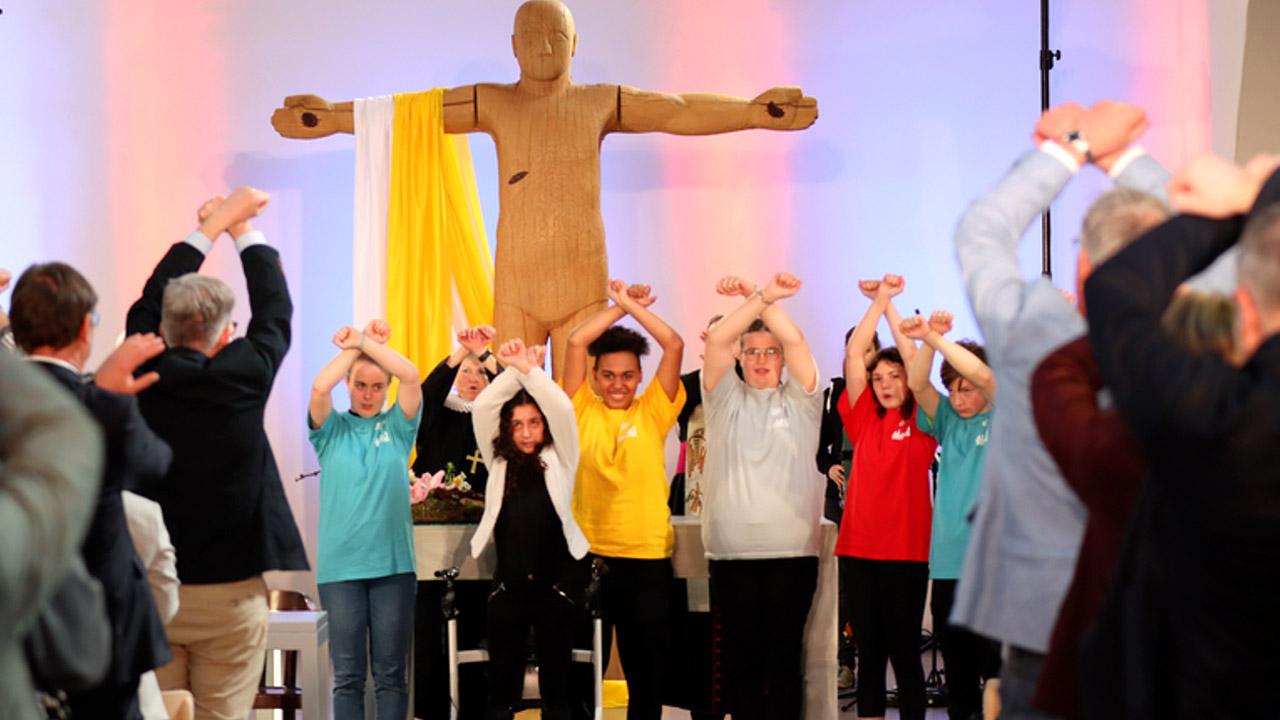 Junge Leute heben die Arme in die Höhe, dahinter eine große, hölzerne Jeusus-Statue